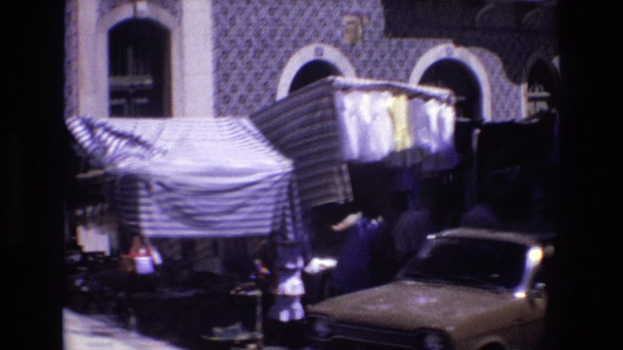 1972 Portugal street-market view with striped canvas covers, hanging fabric, pedestrians near stalls, tiled building surfaces, arched windows, and a classic car at right.