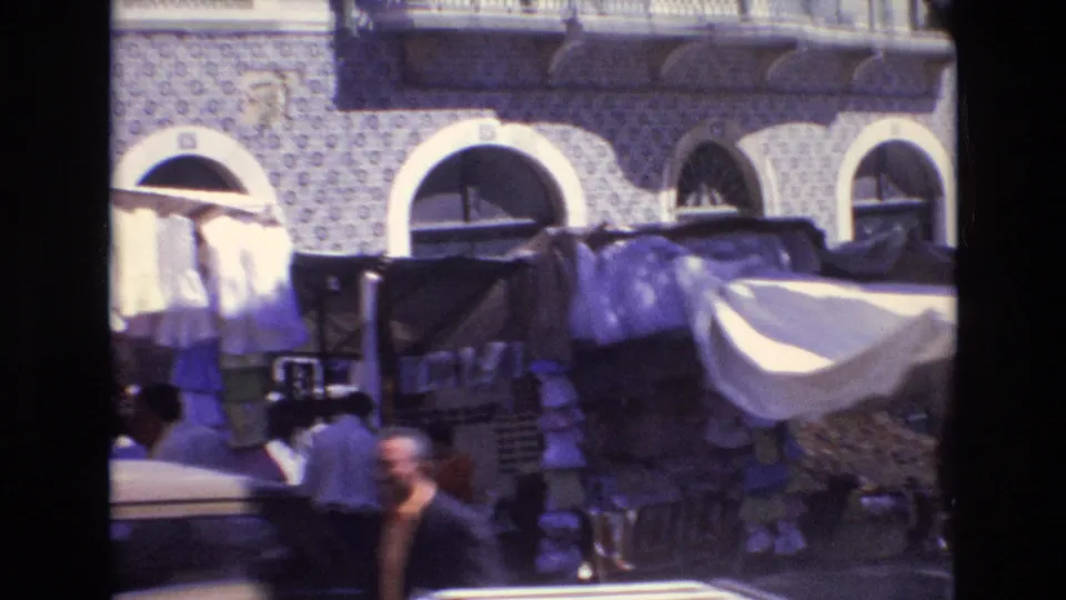 1972 Portugal home-movie frame of a street market below arched windows and patterned tilework, with fabric-covered stalls and blurred pedestrians passing close to camera.