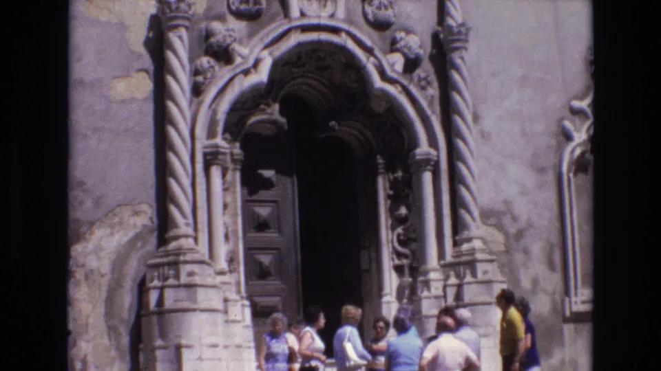 1972 Portugal home-movie frame showing tourists gathered below a massive carved stone entrance with twisted columns, arched ornament, and a dark wooden door.