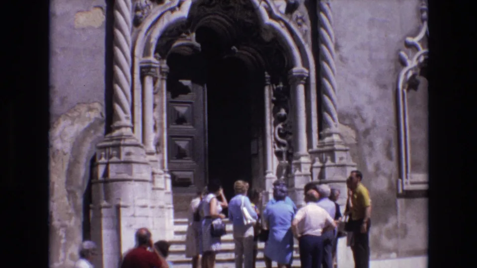 1972 Portugal home-movie frame showing tourists talking near the threshold of a historic building entrance with heavy carved stonework and a dark recessed door.