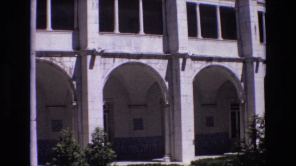 1972 Portugal home-movie frame showing part of a historic building facade with arched lower openings, upper windows, stone columns, and shaded foreground edges.