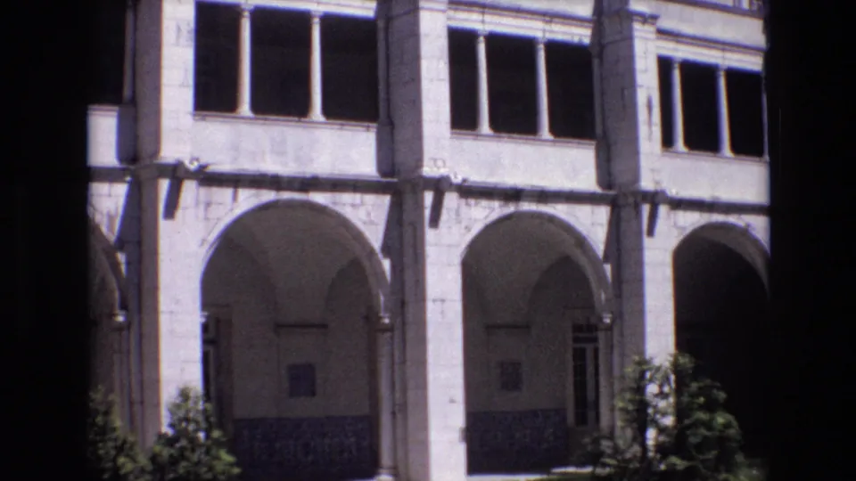 1972 Portugal 8mm frame showing a pale stone courtyard facade with repeated arches, columns, dark window openings, tiled lower walls, and small shrubs.
