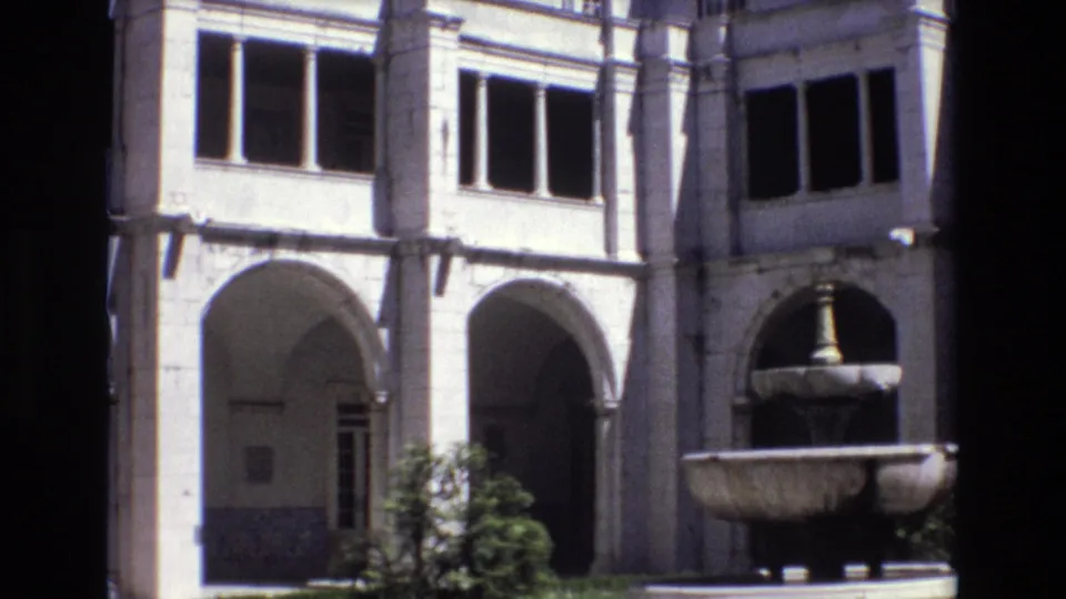 1972 Portugal 8mm frame showing the courtyard corner with pale stone arches, dark upper windows, a planted shrub, and the edge of a stone fountain basin.