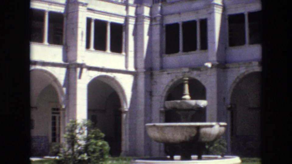 1972 Portugal 8mm frame showing a central stone fountain before a historic courtyard facade with arched openings, windows, tiled wall sections, and garden shrubs.