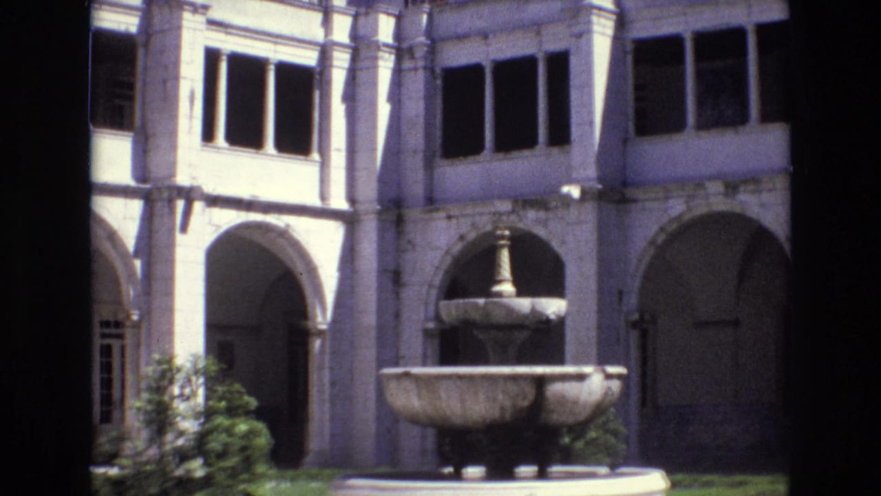 1972 Portugal 8mm frame showing a sunlit pale stone courtyard facade with arched colonnades, dark windows, greenery, and a two-tiered fountain in front.