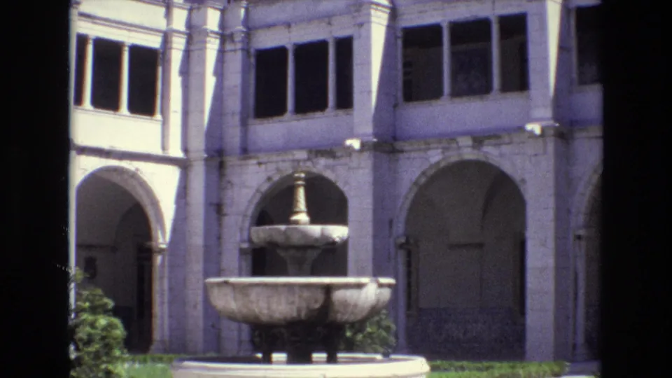 1972 Portugal travel-film frame with a stone fountain centered before a white building, arched walkways, upper windows, and small areas of greenery.