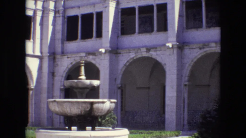 1972 Portugal 8mm frame showing the courtyard fountain lower in the shot, with white stone arches, recessed doors, dark windows, and planted greenery behind it.