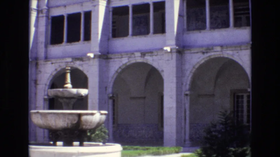 1972 Portugal 8mm frame of a pale courtyard building with repeated arches, multi-paned windows, a stone fountain, shrubs, and strong sun-shadow contrast.