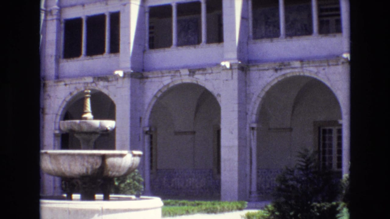 1972 Portugal home-movie frame showing a stone fountain before pale arched walkways, columns, and shadowed upper windows in a historic courtyard.