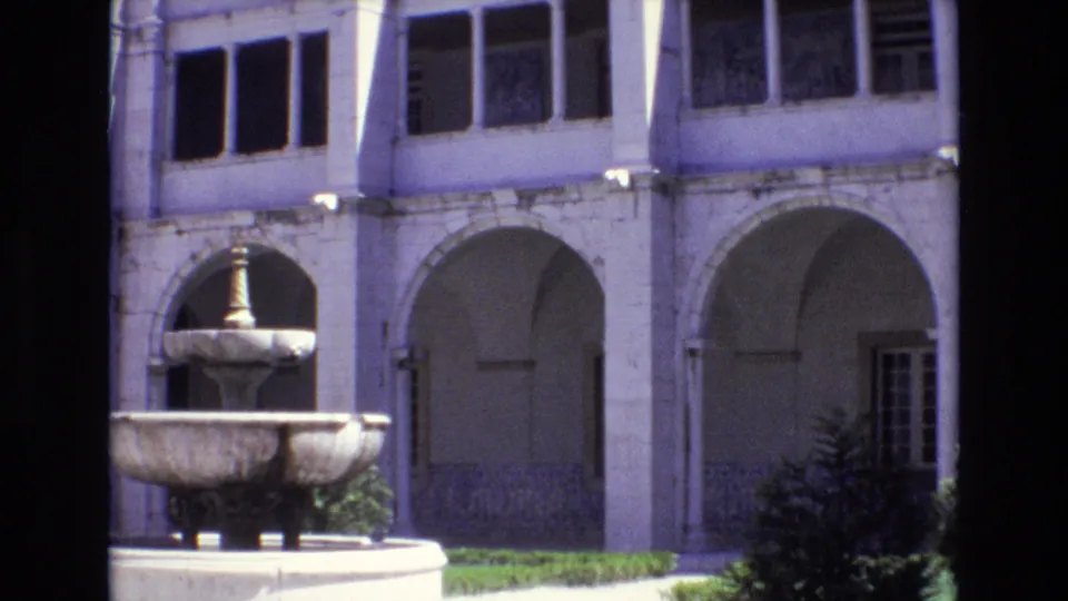 1972 Portugal home-movie frame showing a stone fountain before pale arched walkways, columns, and shadowed upper windows in a historic courtyard.