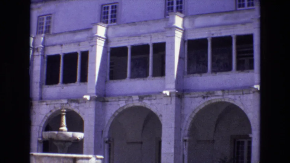 1972 Portugal home-movie still with a fountain at lower left, arched stone walkways, pale columns, upper windows, and a small area of greenery.
