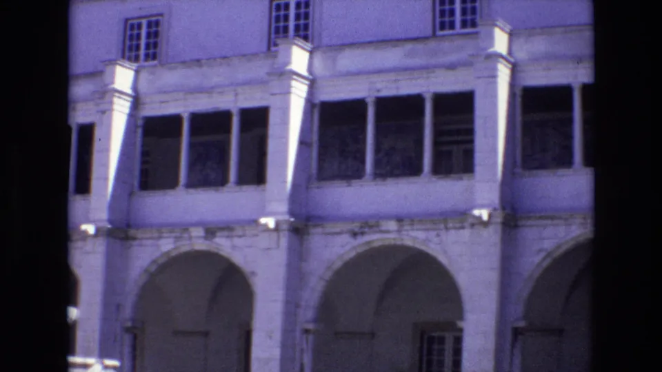 1972 Portugal frame showing the courtyard building from a slightly shifted angle, with arched walkways, tall windows, columns, and part of the fountain at the left edge.