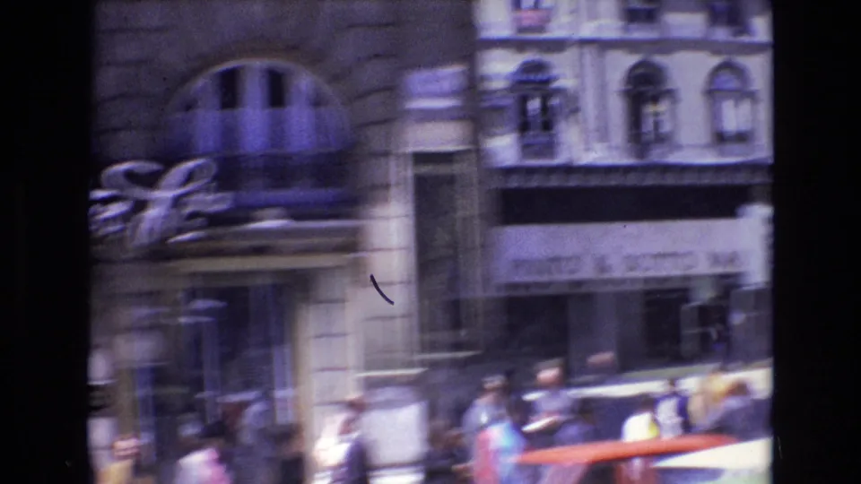 1972 Portugal home-movie street frame showing a moving view of older storefronts, pedestrians on the sidewalk, and vehicles near the curb.