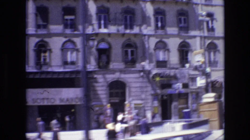 1972 Portugal frame showing pedestrians along a downtown sidewalk below ornate building windows and partially readable storefront signage.