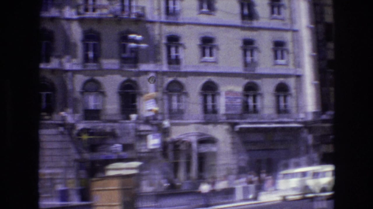 1972 Portugal 8mm frame showing a multi-story urban facade, sidewalk pedestrians, storefront awnings, and passing cars in a busy central street.