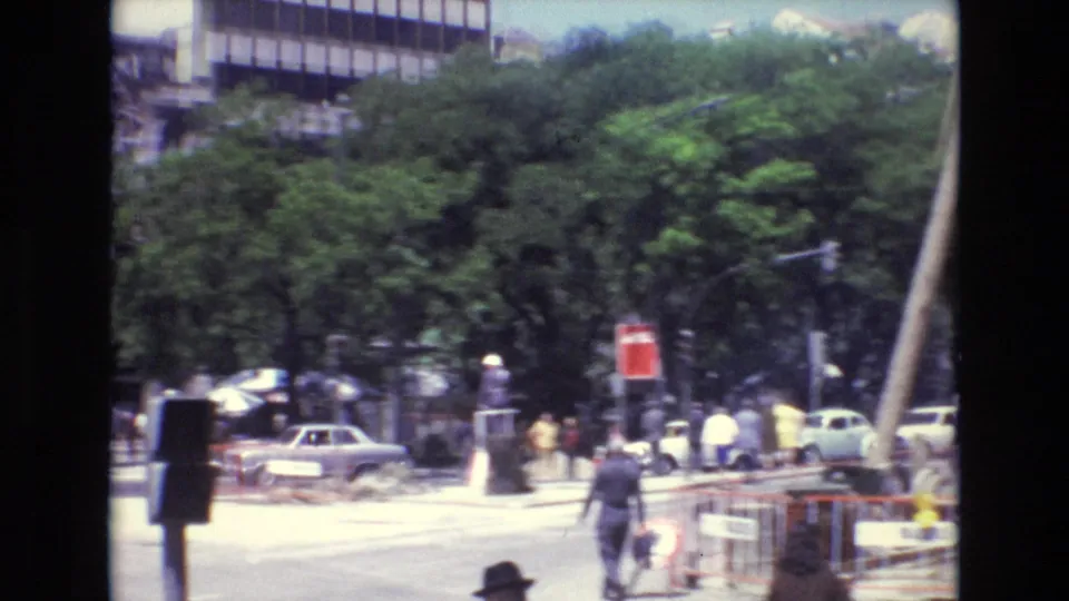 1972 Portugal urban intersection with pedestrians crossing near cars, leafy trees, construction barriers, and a red sign or signal in the street.