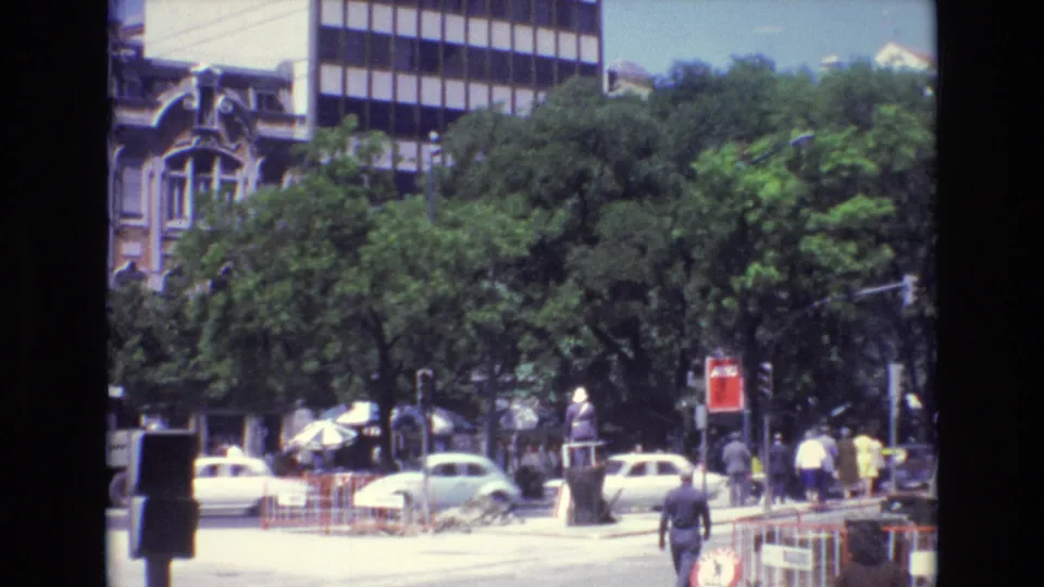 1972 Portugal street view with cars moving past pedestrians, dense green trees, construction barriers, older architecture, and a modern office block in the background.