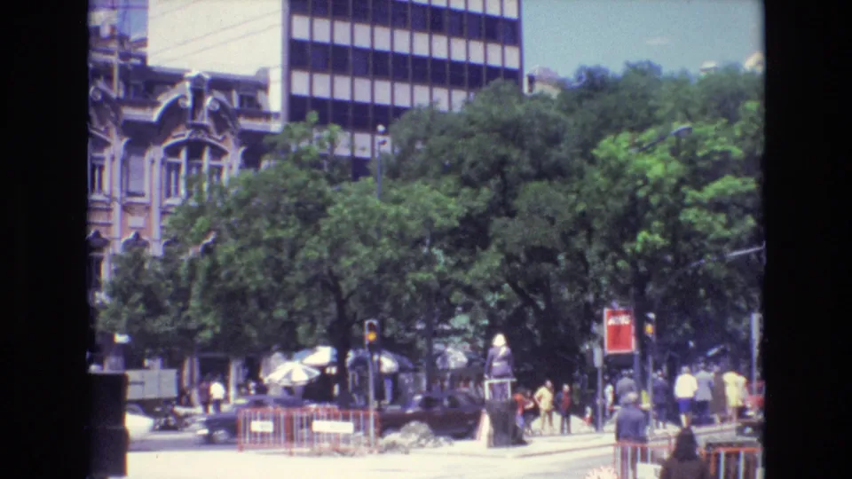 1972 Portugal city corner with pedestrians near construction barriers, cars on the roadway, leafy trees, older facades, and a modern high-rise beyond.