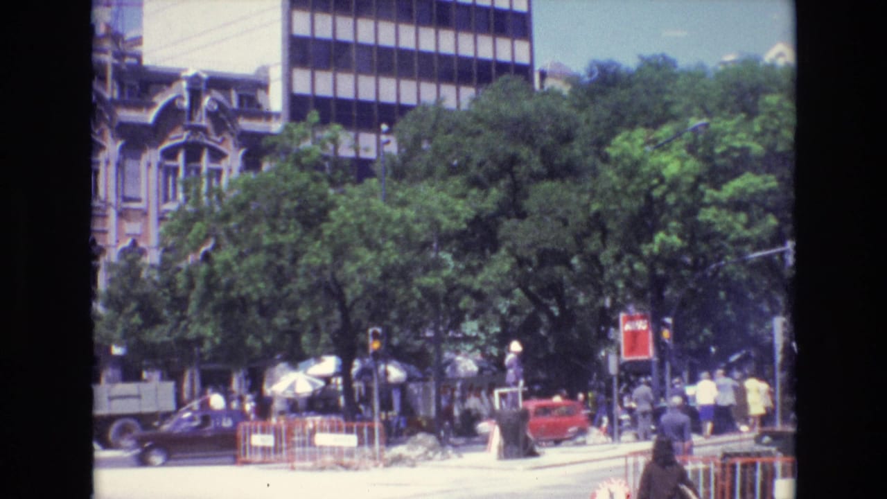 1972 Portugal home-movie frame showing a busy tree-lined city street with pedestrians, classic cars, traffic signals, umbrellas, and older architecture beside a modern facade.