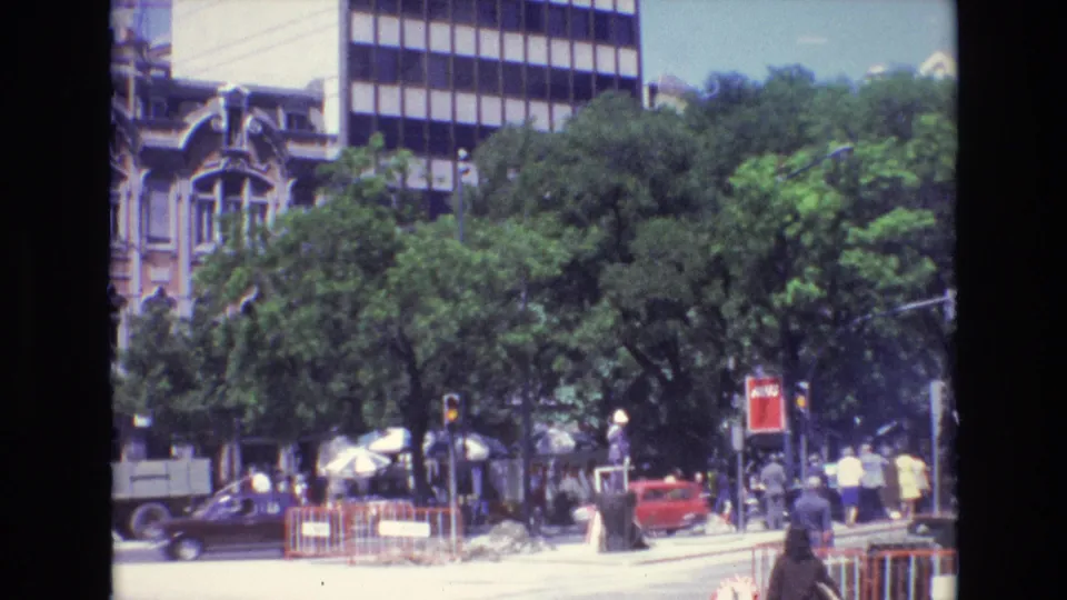1972 Portugal home-movie frame showing a busy tree-lined city street with pedestrians, classic cars, traffic signals, umbrellas, and older architecture beside a modern facade.