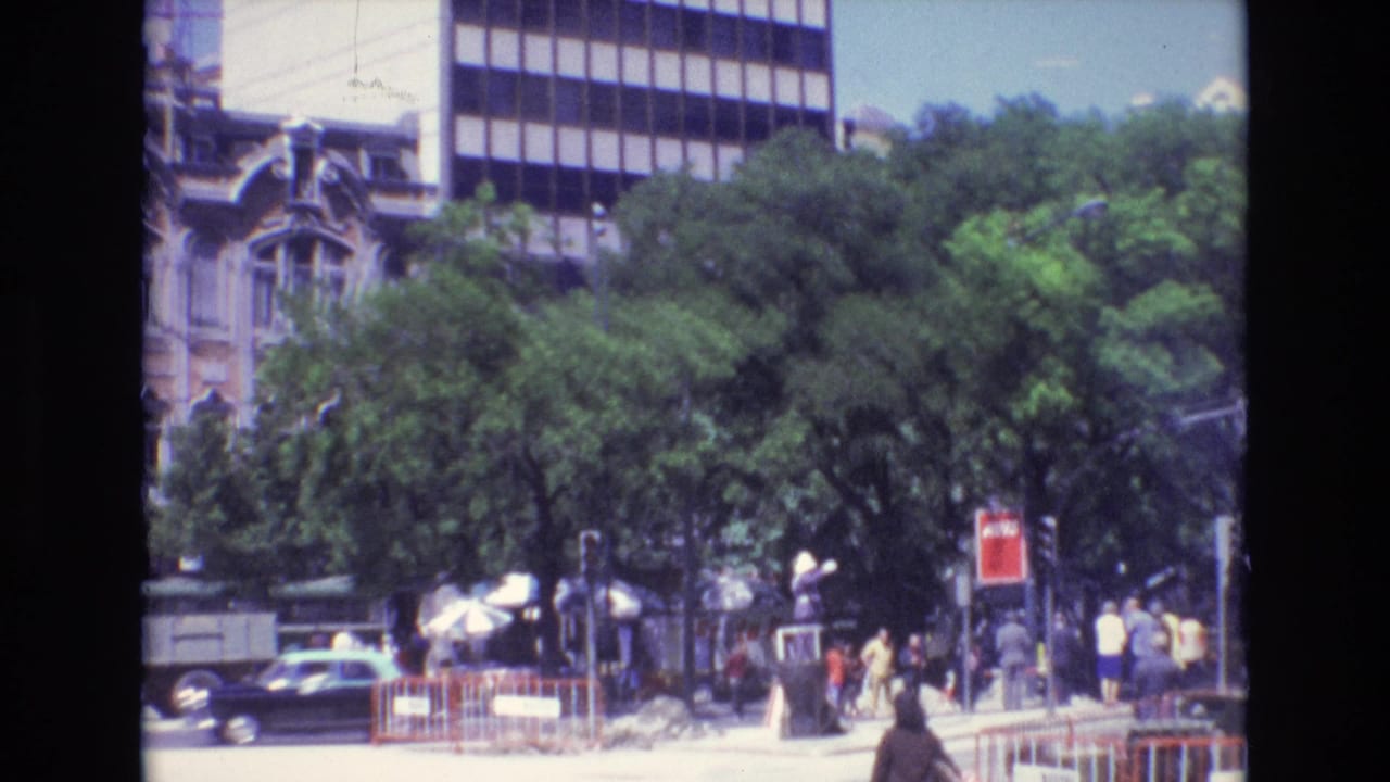 1972 Portugal street view with people walking and gathering near trees, cars, traffic lights, a red sign, umbrellas, and contrasting historic and modern building fronts.