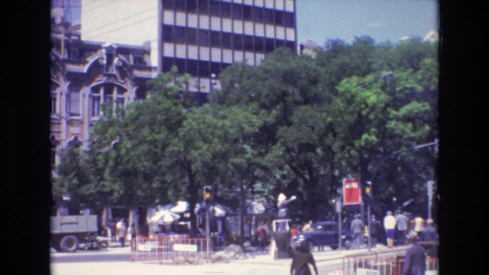 1972 Portugal home-movie view of a city street with leafy trees, pedestrians, cars, traffic signals, umbrellas, railings, and older architecture next to a modern building.