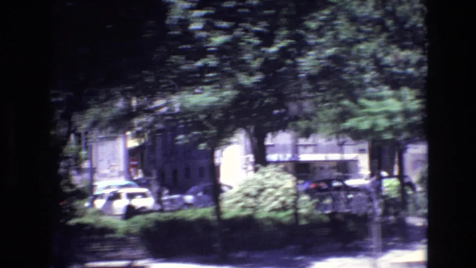 Motion-softened 1972 Portugal urban frame with parked cars, leafy trees, shrubs, and apartment or townhouse facades along the road.