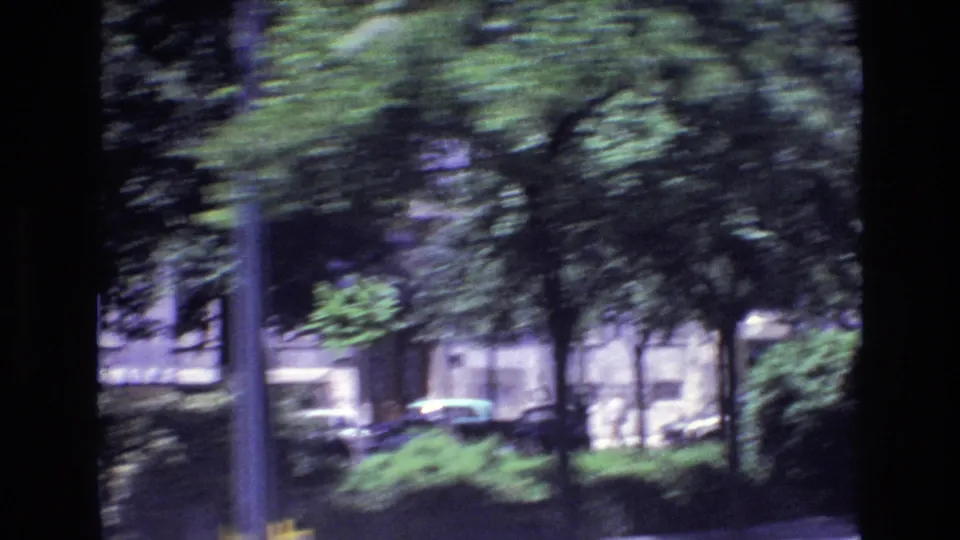 1972 Portugal driving-view frame with a street pole, roadside trees, low greenery, cars, and pale building facades passing in blur.