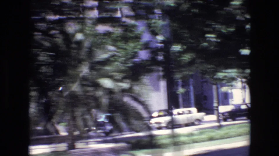 1972 Portugal 8mm driving view with mature trees, parked classic cars, grass, and building facades along an urban street.
