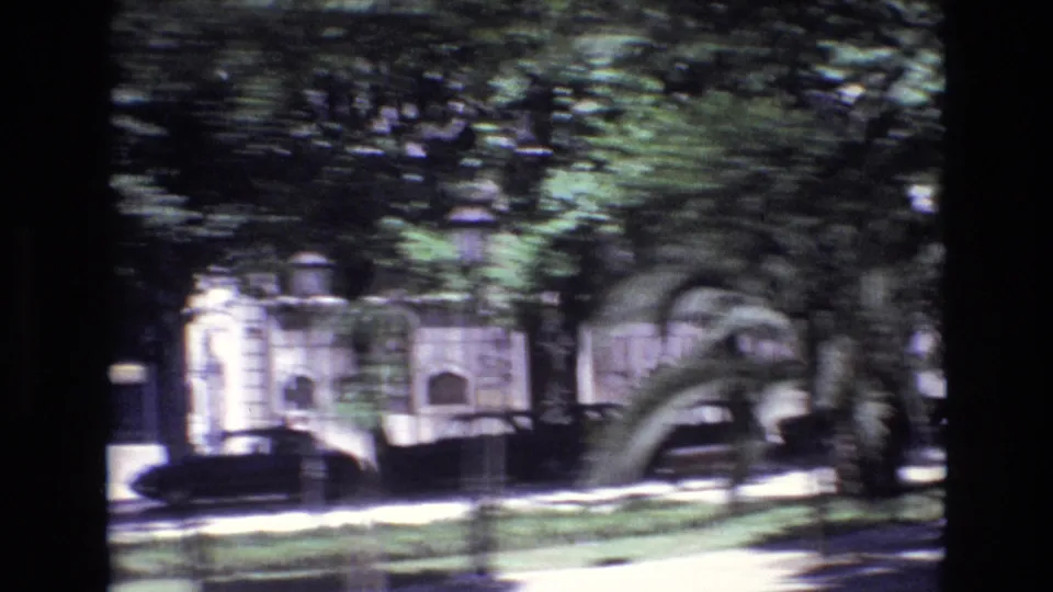 1972 Portugal frame showing a shaded tree-lined street, parked car, planted verge, and residential facade with arched architectural details.