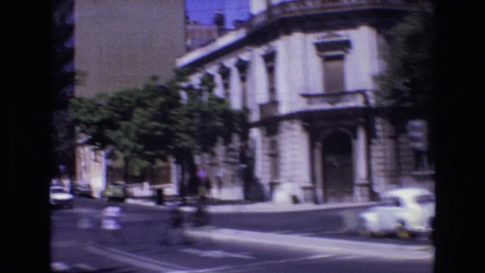 Portugal 1972 street view of a masonry corner building with tall windows, an arched entrance, parked cars, and a sunlit road captured from a moving vehicle.