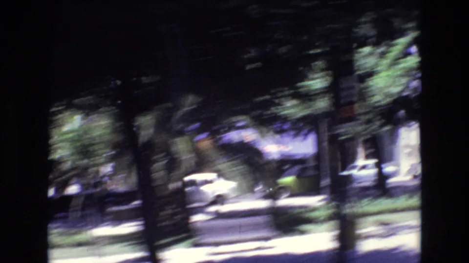 A 1972 Portugal home-movie frame shows a shaded urban roadside with trees, parked or passing cars, building windows, and strong motion blur from travel.