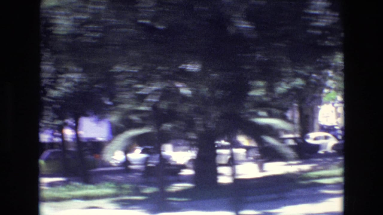 A blurred 1972 Portugal street view with trees, bright daylight, parked cars, and a dark film edge from a moving 8mm camera.