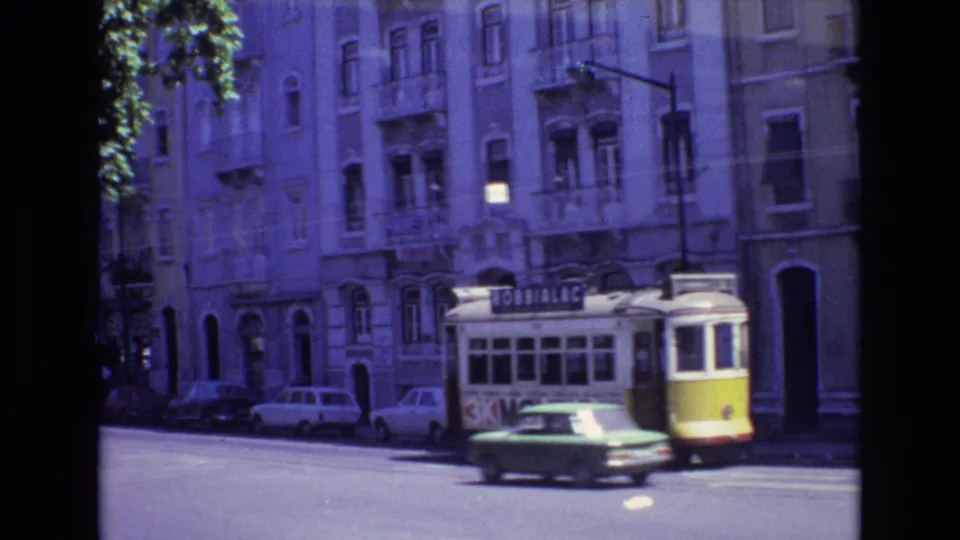 A 1972 Portugal city street with a yellow-and-white trolley, older cars, apartment facades, street trees, and visible trolley signage.