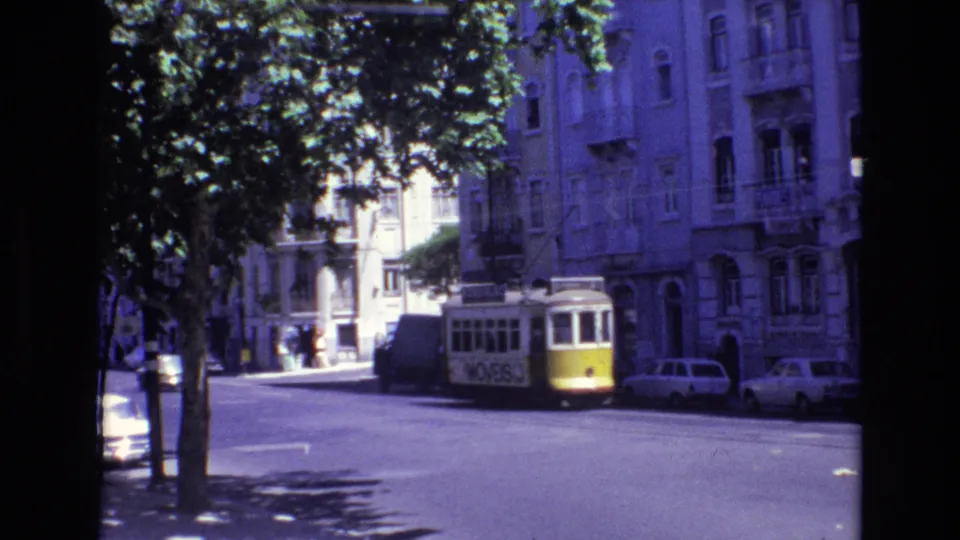 1972 Portugal 8mm frame with a trolley moving away along a shaded street lined with parked cars, trees, and apartment facades.