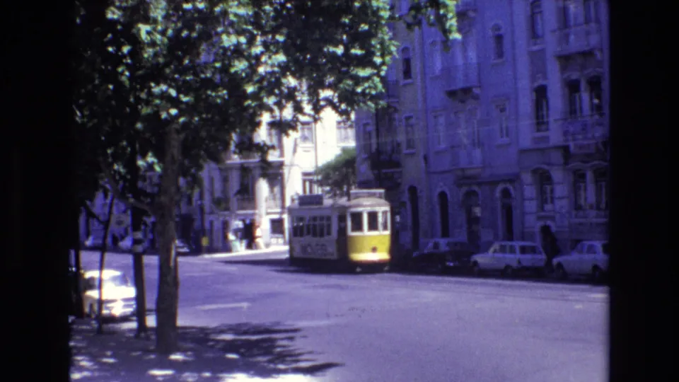 1972 Portugal city street frame showing the trolley farther from camera, with parked cars, tree shade, and residential building fronts in view.