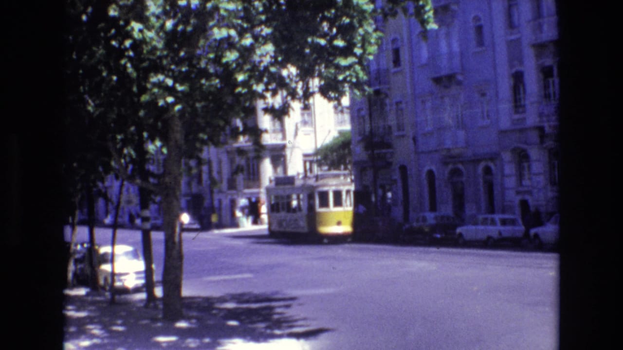1972 Portugal street view from 8mm footage, showing a yellow-and-white tram approaching along a sunlit urban road with trees, parked cars, and older buildings.