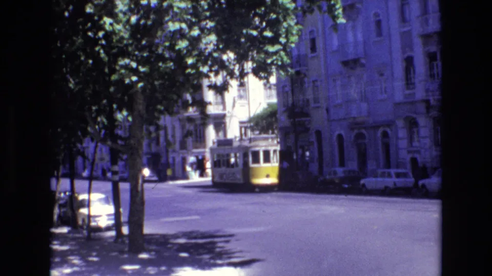 1972 Portugal street view from 8mm footage, showing a yellow-and-white tram approaching along a sunlit urban road with trees, parked cars, and older buildings.