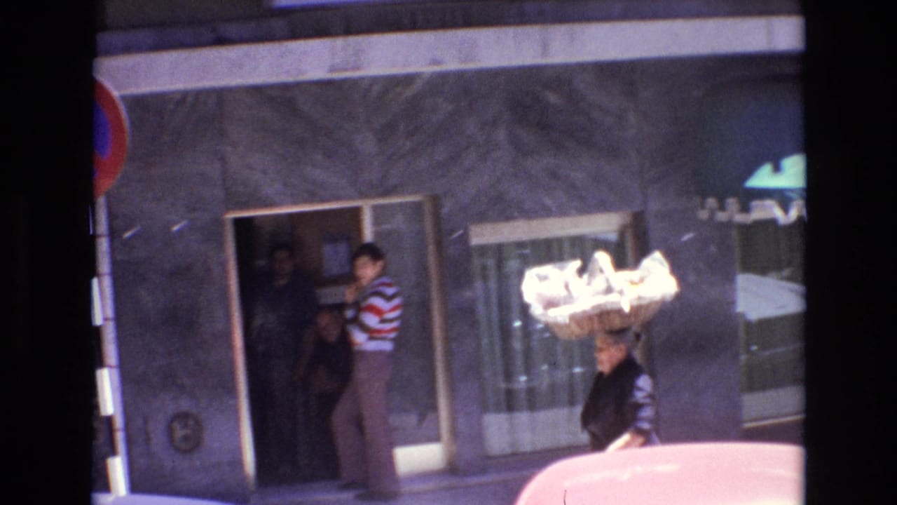 1972 Portugal street scene with a woman in dark clothing carrying a large woven basket on her head near a gray storefront and a man in a striped sweater.