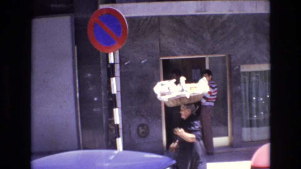 1972 Portugal 8mm frame showing a woman walking with a large basket balanced on her head beside a no-parking sign, storefront doorway, and parked car roof.