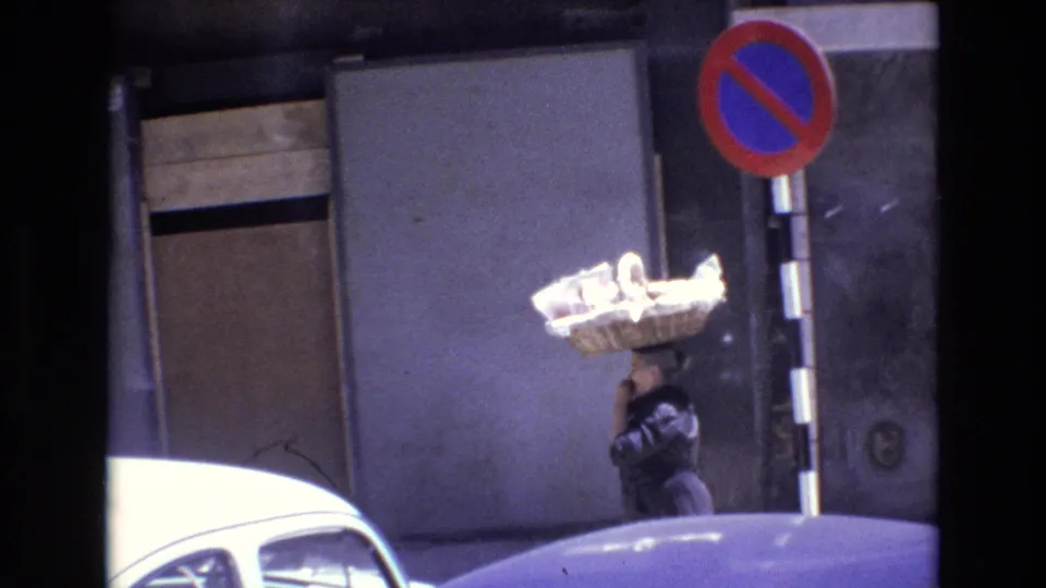 1972 Portugal street footage frame with the woman continuing past a gray building, balancing a large woven basket on her head near a no-parking sign and parked cars.