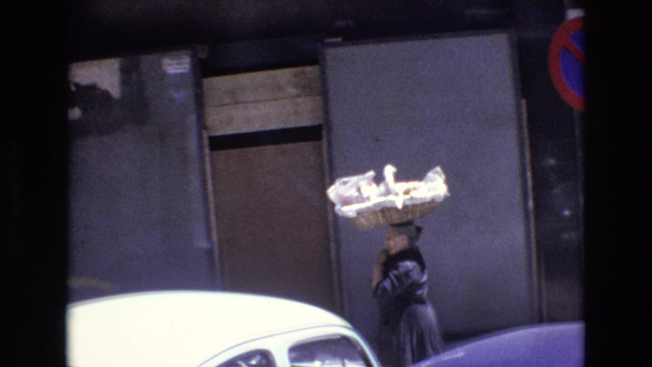 Woman balancing a large basket on her head beside parked cars on a city street.