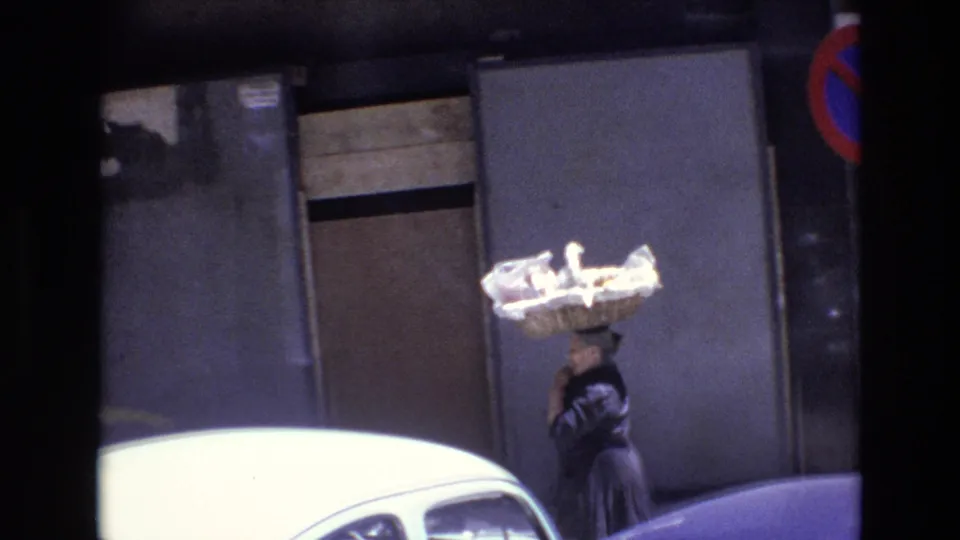 In Portugal in 1972, a woman in dark clothing balances a large light-colored basket on her head while walking past parked cars and a no-parking sign on an urban street.