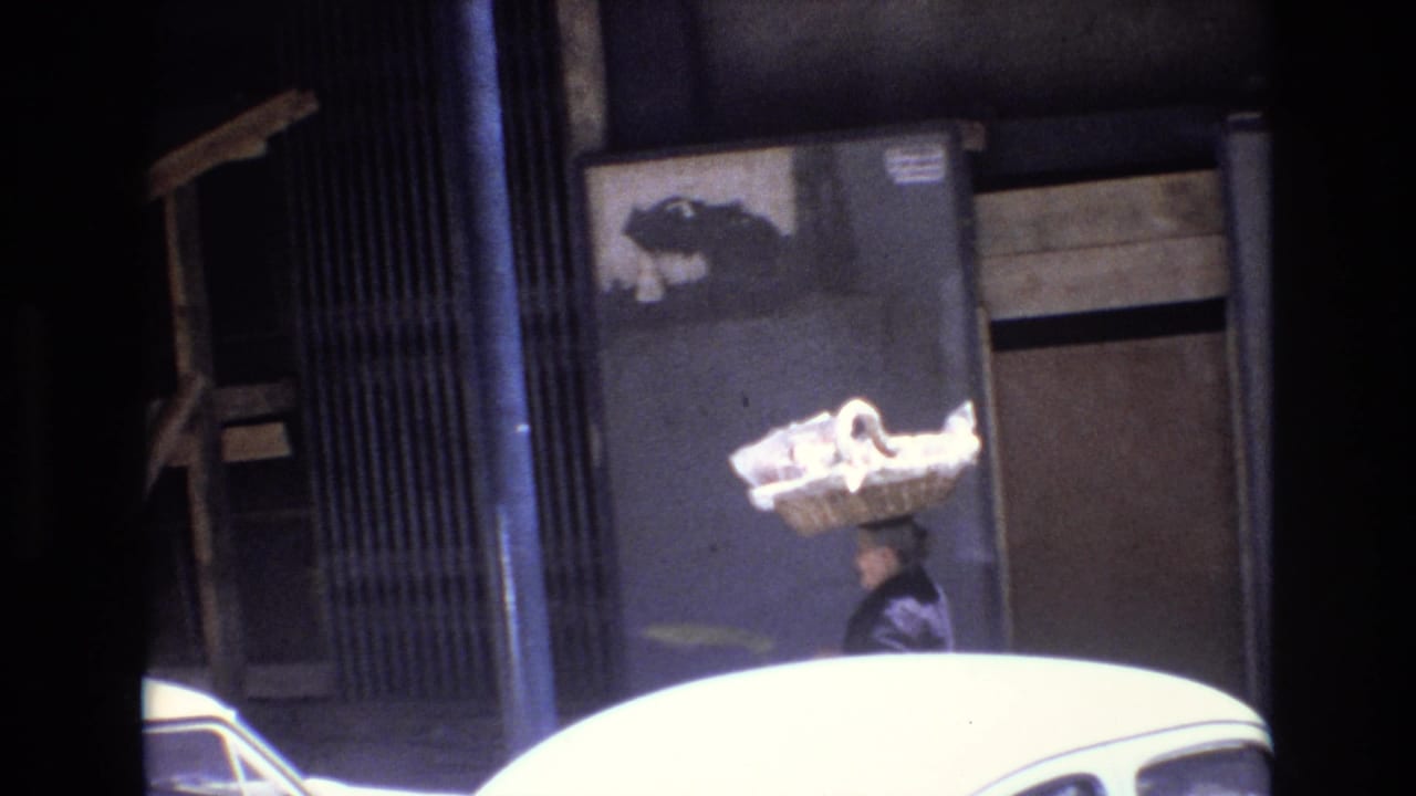 Woman with a basket on her head passes a parked white car and building entrance.