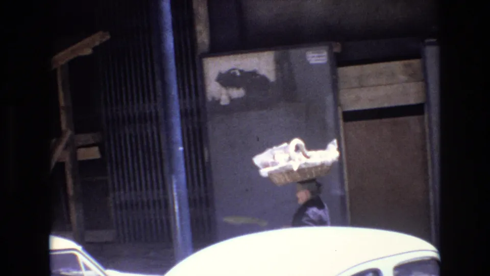 The 1972 Portugal street view shows the woman continuing along the sidewalk with a large basket balanced on her head, partly obscured by parked cars and dark film-frame borders.