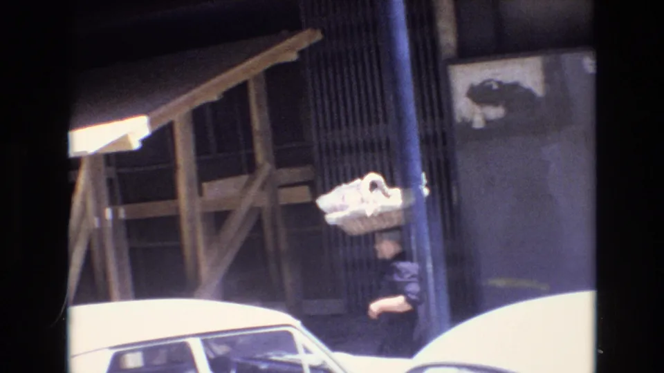A handheld 8mm frame from Portugal in 1972 catches the woman with the basket near a parked white car, a gray wall, and wooden construction-like framing by the sidewalk.