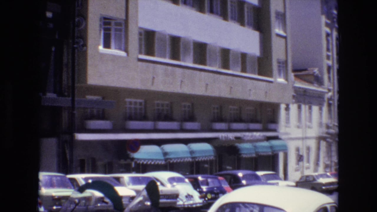1972 Portugal home-movie frame showing curbside parked cars, a beige multi-story building, teal awnings, and a traffic sign along an urban street.