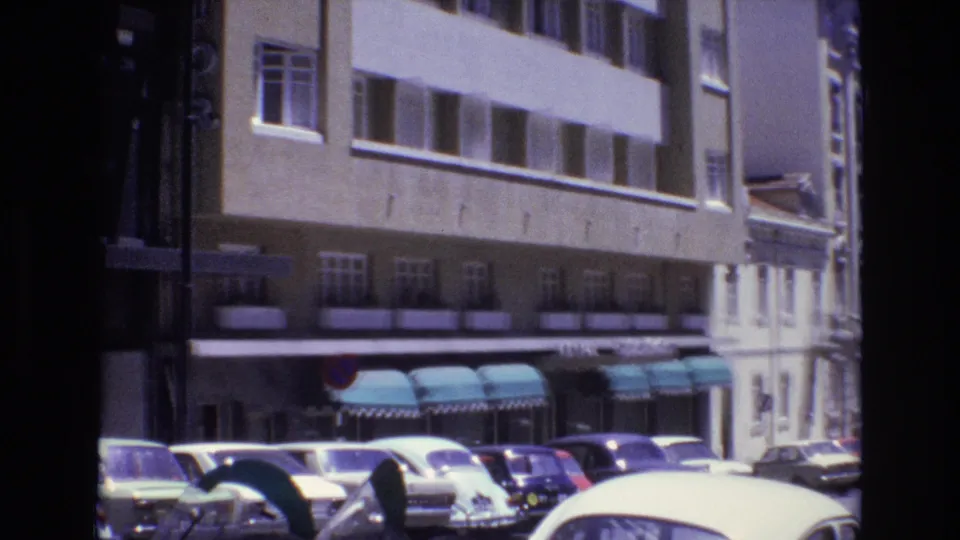 1972 Portugal home-movie frame showing curbside parked cars, a beige multi-story building, teal awnings, and a traffic sign along an urban street.