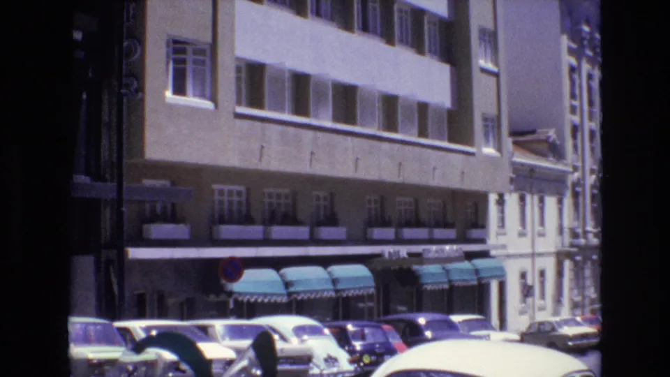 1972 Portugal street view with several parked 1970s cars, a large beige facade, window rows, teal awnings, and a red-and-blue traffic sign.