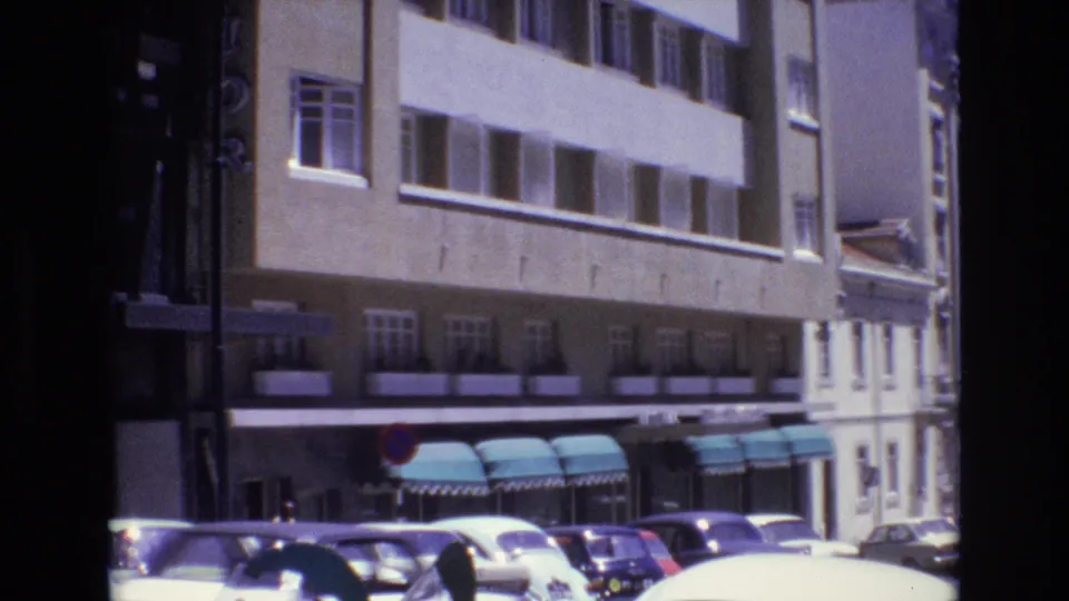1972 Portugal home-movie frame showing classic parked cars, a beige commercial-looking building frontage, teal awnings, and soft 8mm film grain.